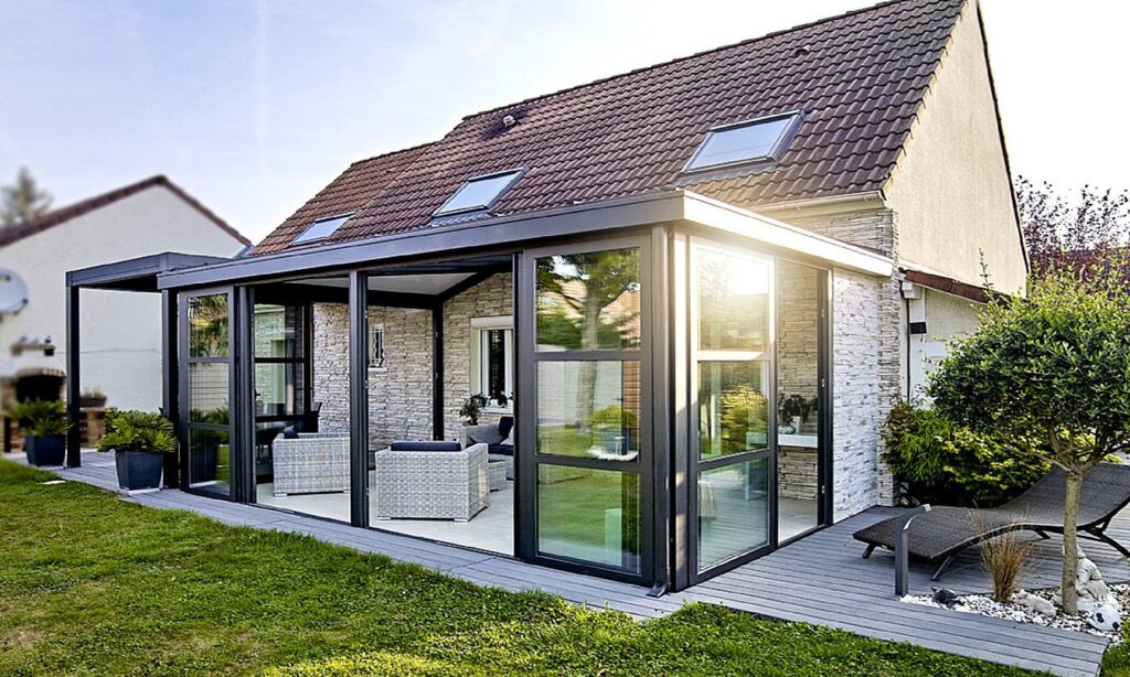 Extension véranda sur une maison en pierre, mobilier en osier sur terrasse en bois, lumière du soir et velux sur le toit.