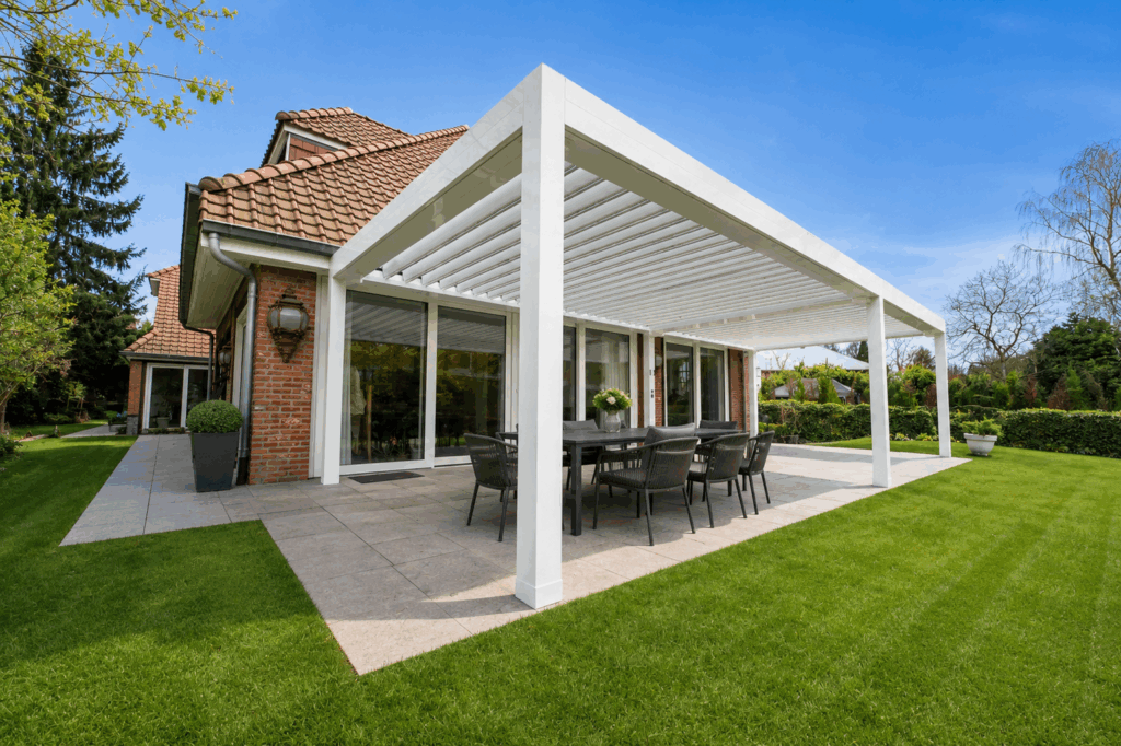 Pergola bioclimatique blanche attenante à une maison en briques à toiture en tuiles, table à manger noire sur terrasse et pelouse.