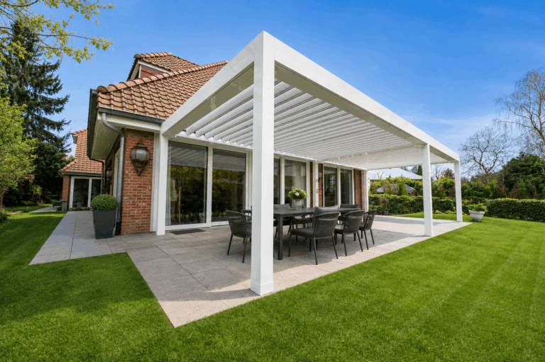 Pergola bioclimatique blanche attenante à une maison en briques à toiture en tuiles, table à manger noire sur terrasse et pelouse.
