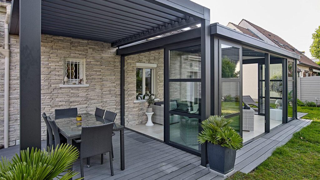 Pergola et véranda attenantes à une maison en pierre, mobilier de jardin en osier sur terrasse en bois.