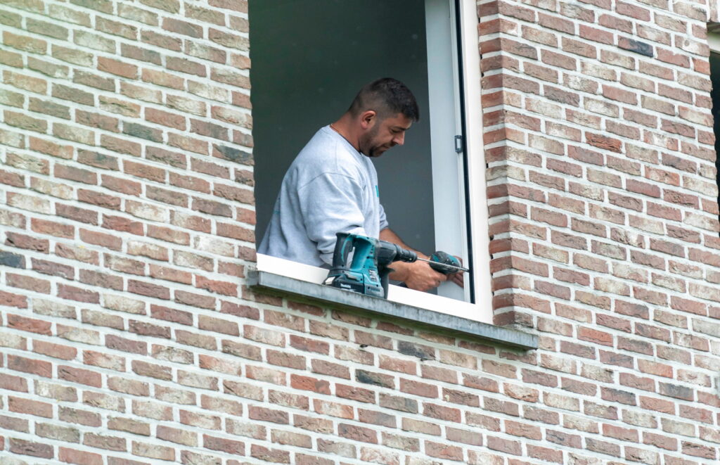 Technicien Abihome perçant un châssis depuis l'extérieur d'un immeuble en briques, à l'étage.