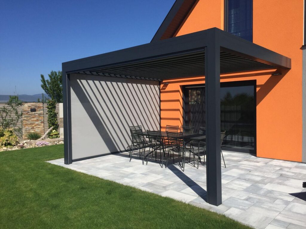 Pergola bioclimatique anthracite avec écran latéral rétractable, terrasse en pierre devant une maison orange, table à manger en métal.