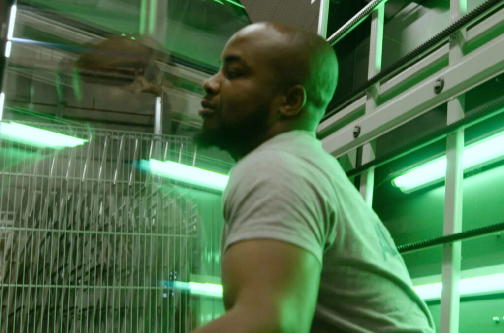 Technician working under green lighting in a production workshop, near a metal cage and industrial rails.