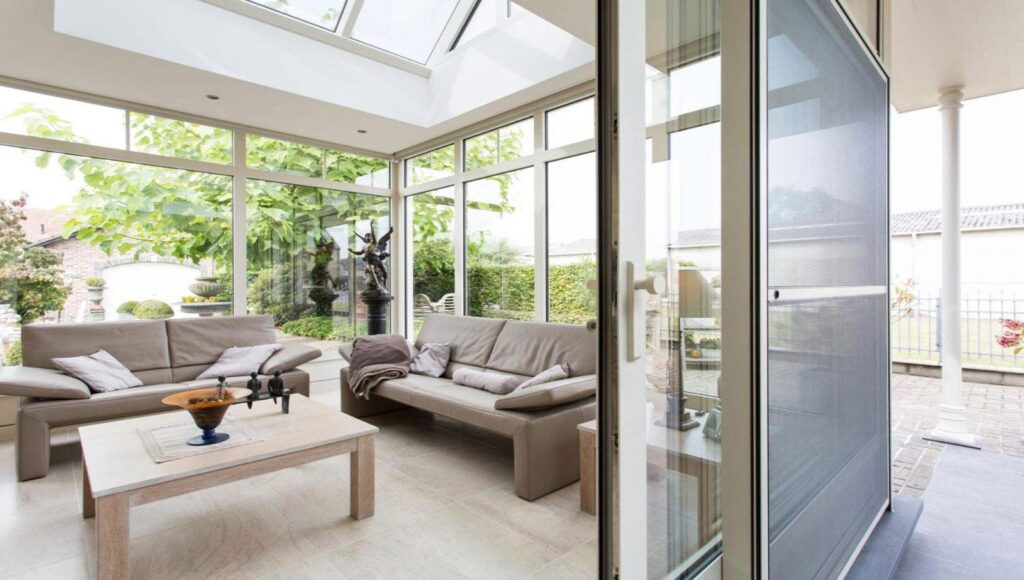Intérieur d'une véranda à toiture plate avec velux, canapés en cuir beige et vue sur le jardin.