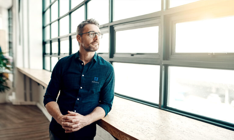 Collaborateur Abihome en chemise à logo, debout près d'une grande fenêtre industrielle dans un espace de bureau lumineux.