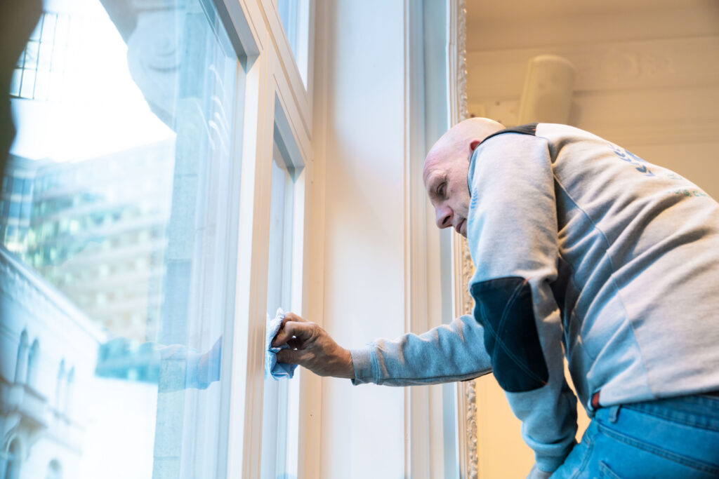 Technicien Abihome nettoyant un châssis en bois depuis l'intérieur, vue sur la ville à travers la fenêtre.
