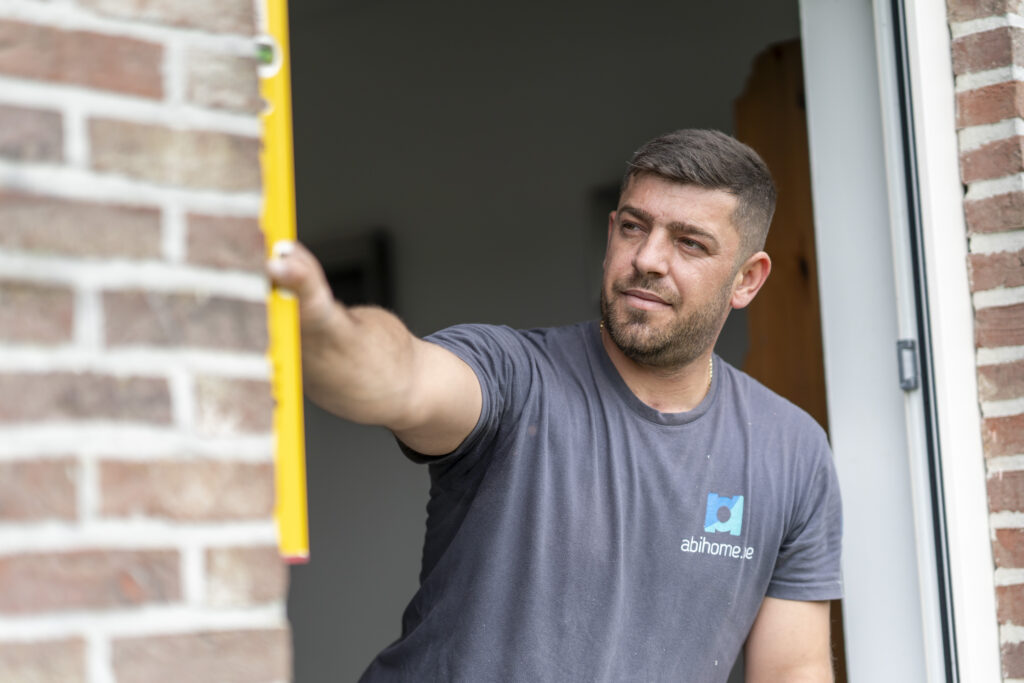 Technicien Abihome vérifiant l'aplomb d'un dormant de porte à l'aide d'un niveau, entrée d'une maison en briques.
