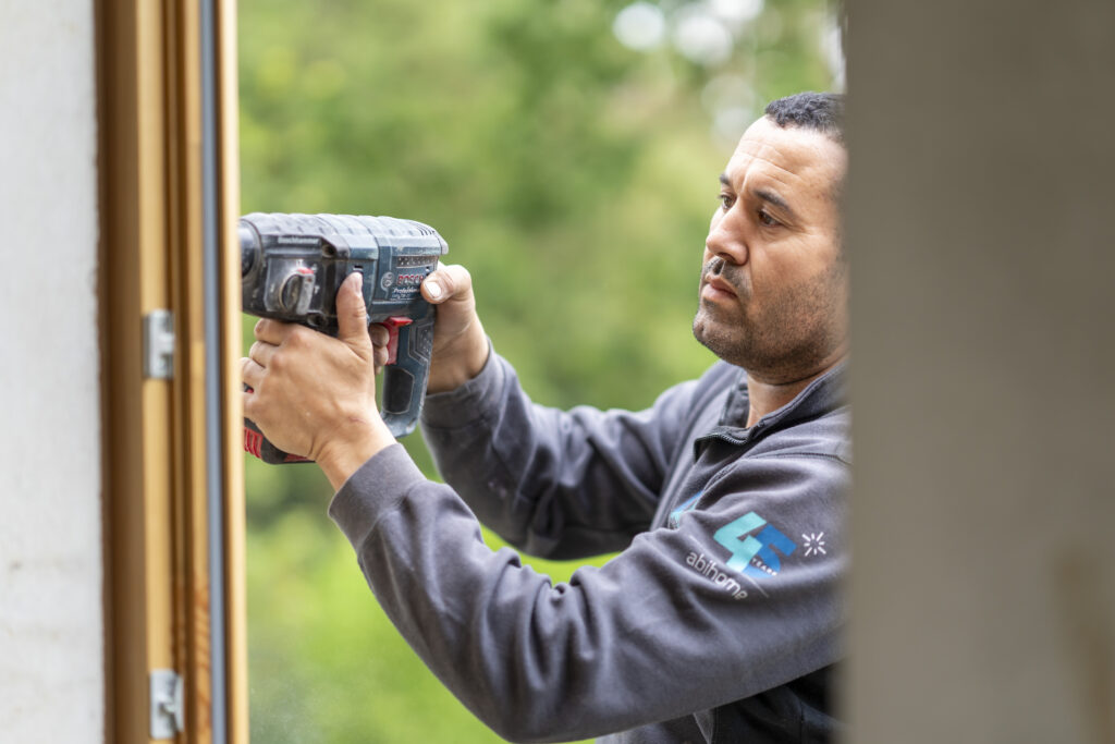 Technicien Abihome perçant un dormant de porte ou châssis à l'aide d'une perceuse, environnement extérieur verdoyant.