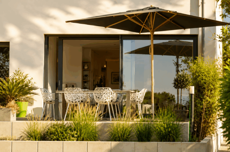 Terrasse ombragée par un grand parasol, baie coulissante PVC Premidoor76 ouverte sur un intérieur avec table en bois et chaises en osier.