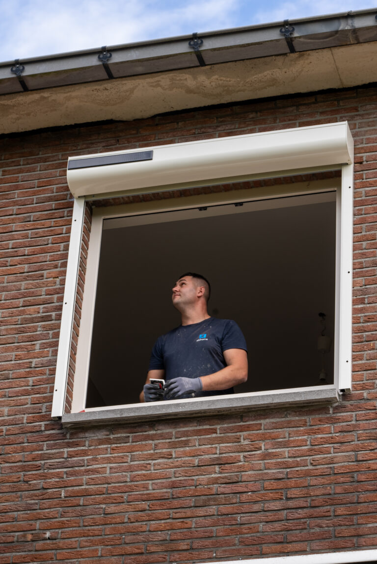 Technicien Abihome installant un coffre de volet roulant solaire au-dessus d'une fenêtre, façade en briques.