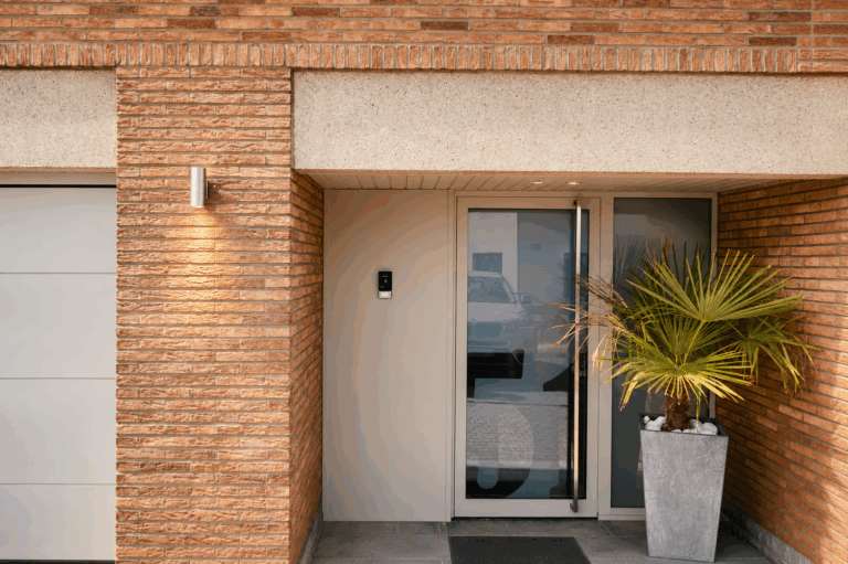 Entrée d'une maison en briques avec porte vitrée en aluminium à imposte latérale, porte de garage en bois à côté, palmier en pot et interphone.