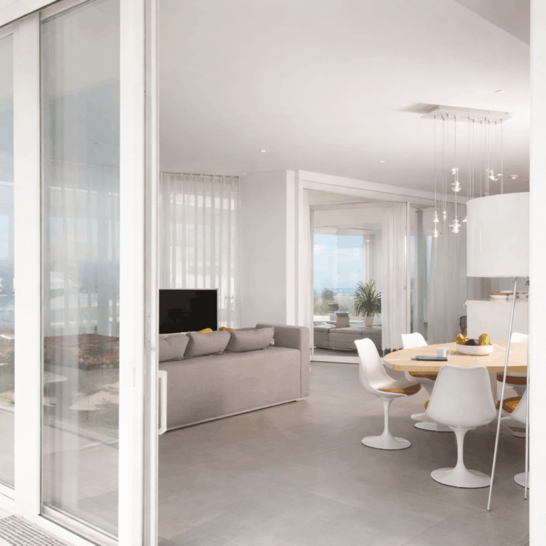 Baie coulissante Premidoor76 ouverte sur un intérieur lumineux avec table à manger, chaises blanches et vue dégagée sur la mer en arrière-plan.