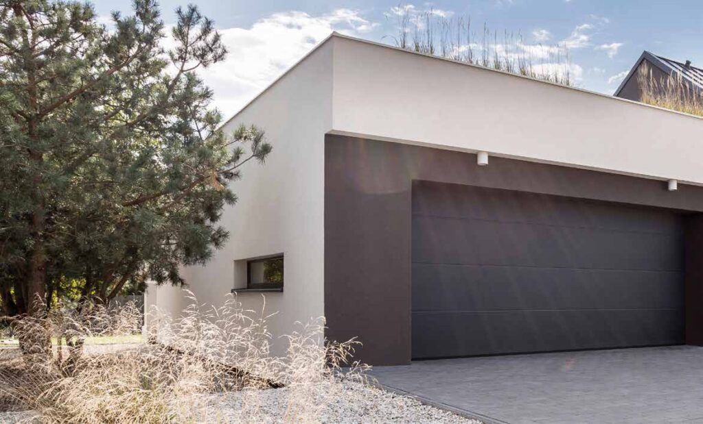 Porte de garage sectionnelle plafond gris anthracite sur une maison contemporaine, pin et allée en gravier.