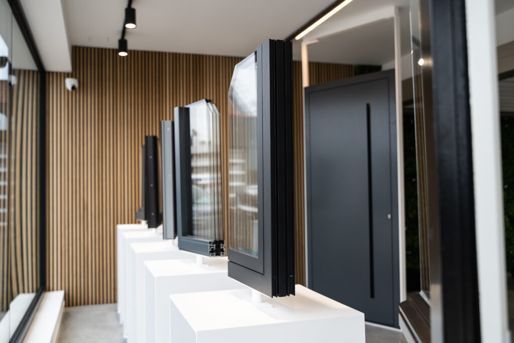Showroom Abihome avec échantillons de châssis et porte d'entrée noire exposés sur socles blancs, mur en bois clair.