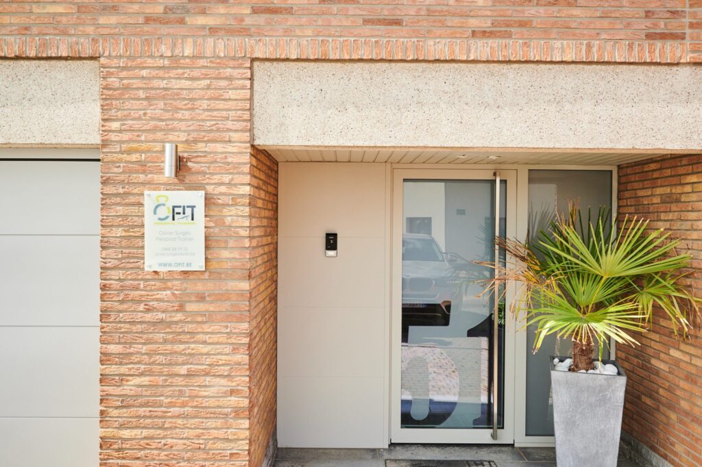 Entrée d'une maison en briques avec porte d'entrée en aluminium blanc cassé, panneau vitré latéral, interphone et palmier en pot.